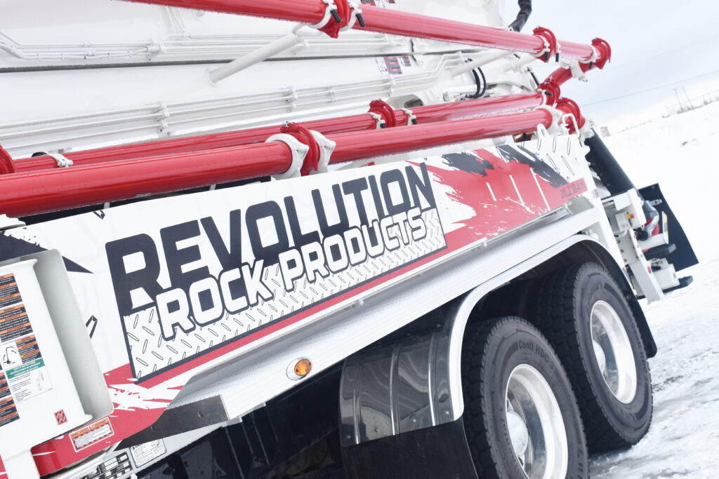 Detail view of the side of one of Revolution Rock Products' trucks, a white and red concrete pumping truck with a large white boom arm with red piping. The side of the truck features a large colourful decal with the Revolution Rock Products logo and splashes of red and black against a checkerplate background.
