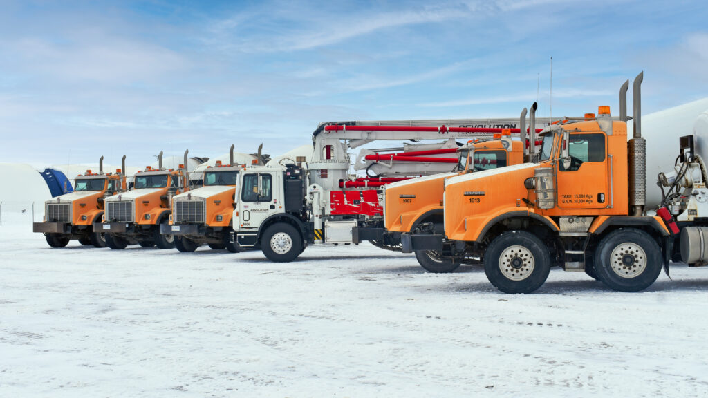 Lineup of six Revolution Rock Products trucks, including five orange concrete mixer trucks and one white and red concrete pumping truck in the middle with a large white boom arm with red piping.