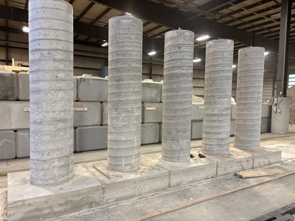 A line-up of five large precast concrete columns with thick square bases in the Revolution Rock Products shop. In the background can be seen large concrete blocks, the kind used to construct barriers for large scale construction projects.